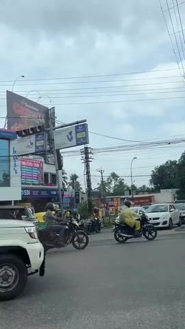 The video captures a street scene during daytime, under an overcast sky, in what appears to be a bustling commercial area. The camera seems to be in a moving vehicle, panning across the road and roadside establishments.

Several vehicles are visible in motion. A black Kia Carens SUV moves from left to right, followed by a yellow Piaggio Ape auto-rickshaw. A truck with a blue tarp passes on the right side of the frame initially. Later, a white truck with metal railings, bearing the license plate "KL 63 J 2187" (indicating Kerala, India), is seen. A red Maruti Suzuki WagonR car also drives past from left to right. Other vehicles, including a white car, are visible in the background.

The scene features multiple buildings and commercial signs. A prominent HDFC Bank branch is visible, displaying its name in both English and Malayalam script. Adjacent to it, a dark building features a large circular yellow sign for "let's EAT. Bakers." Another shop sign with "kair" written in Malayalam is also visible, along with partially visible text "RYTEL MEDICALS".

Overhead, several large billboards and traffic lights are present. One billboard advertises "Dominos Chick 'n' Chizza" for "$299". Another large billboard promotes "Mentor ET-ENGINEERING COLLEGES" with the slogan "LEARN BEYOND BOOKS" and a phone number "+91 94959 55111". A dense network of overhead power lines and cables crisscrosses the sky. The general activity is typical urban traffic flow on a busy road, with vehicles moving in various directions. No specific people are clearly identifiable.