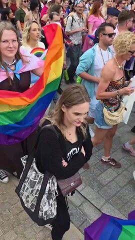 A large crowd of people are walking down a street, many of them holding or wearing rainbow flags, indicating a pride parade. The people are diverse in age and gender, and many are dressed in casual clothing. Some are smiling and looking at the camera, while others are looking forward or interacting with each other. The street is paved, and there are buildings on either side. The weather appears to be sunny.
