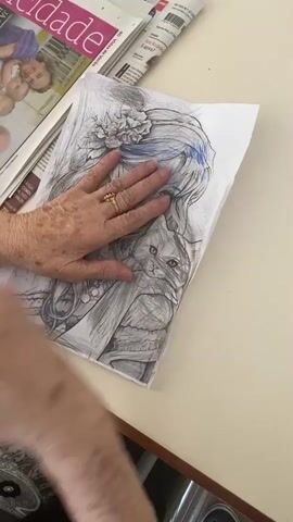 A close-up shot shows an elderly person's hands coloring a drawing of a woman with long, flowing hair and large eyes. The drawing is on a white sheet of paper and is placed on a table next to a stack of magazines. The person is using a dark-colored pencil to color the woman's hair with blue streaks. The person's left hand rests on the drawing, holding it in place, while their right hand holds the pencil and carefully colors. The person is wearing a gold ring on their left ring finger. The background is blurred, but appears to be an indoor setting with a wooden table and part of a chair visible.