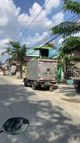 The video shows a street scene from the perspective of someone riding a motorcycle. The road is paved and appears to be in a somewhat urban or semi-urban area. Along the sides of the road, there are buildings, some of which appear to be shops or businesses with signs. One sign clearly visible says "vivo." There are also trees and utility poles with many wires. A yellow truck is parked on the right side of the road. Several motorcycles are visible, including the one the video is being recorded from, and others are being ridden on the street. The sky is blue with some white clouds. The overall impression is a typical day in a street with local commerce and traffic.