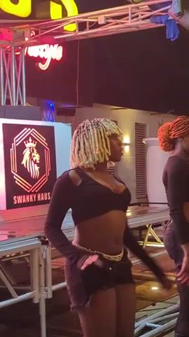 The video opens with a wide shot of a nightclub setting at night. In the foreground, a woman with blonde braided hair and a black crop top and shorts is dancing. She is wearing a gold chain around her waist. To her right, another woman with orange braided hair in a black outfit is also dancing. Behind the first woman, a lit sign displays a lion logo with the text "SWANKY HAUS." In the background, there are more lights and a building structure. The camera zooms in slightly on the dancing women. The overall atmosphere appears to be lively and energetic.