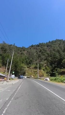 The video shows a view from a moving vehicle on a paved road that curves to the left. The road is flanked by a steep, tree-covered mountainside on the right and a drop-off with a guardrail on the left. Telephone poles and wires run along the left side of the road. Several vehicles, including vans and cars, are visible on the road. The sky is clear and blue.