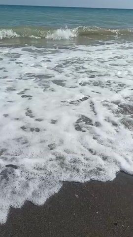 The video shows a beach scene with waves crashing on the shore. The water is blue and clear, and the sky is bright and sunny with a few clouds. There are people walking and playing on the beach in the distance. The sun is shining brightly.