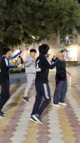 A group of young men are dancing in a public plaza at night. The men are wearing a variety of casual clothing, including matching black tracksuits with a logo on the back. The logo appears to be a stylized image of a dog and the word "ANIMUS." Some of the men are carrying objects, such as a backpack and a colorful ribbon. The plaza is paved with patterned tiles, and there are trees, benches, and lampposts in the background. Some people are sitting or lying down on the benches and ground. The scene is lit by artificial lights. The men are dancing together, moving in a coordinated fashion. The overall atmosphere appears relaxed and social.