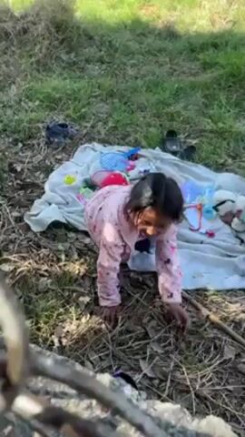 The video shows a young child sitting near a pile of objects spread out on a white cloth. The child is wearing a pink and white patterned shirt and black pants, and has dark hair in braids. The objects on the cloth include children’s toys, books, and accessories. There are pairs of shoes nearby on the grass. The scene appears to be outdoors, in a grassy area with trees in the background. The child is interacting with something on the ground, possibly putting on a pair of shoes.
