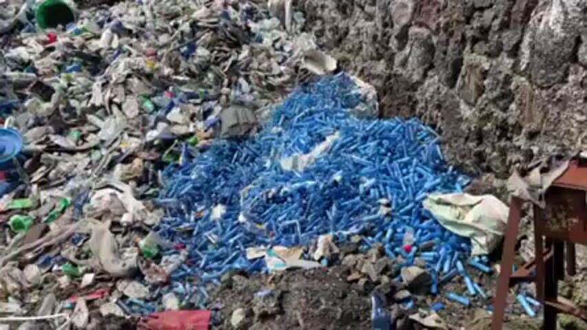 The video shows a large pile of trash and debris including numerous plastic bottles, pieces of plastic, trash bags, buckets, and other miscellaneous waste. The waste is primarily white and grey, but there is a distinct pile of blue plastic objects. The garbage pile is located next to a rock wall and some dirt. A table and chair are also visible.