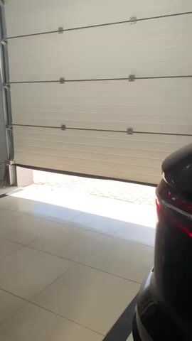 The video shows a man in a suit cleaning the door handle of a silver car in what appears to be a car dealership. The dealership has a clean, tiled floor and a large overhead door is partially open, letting in sunlight. In the background, other cars and a staircase are visible, as are some employees working at desks.