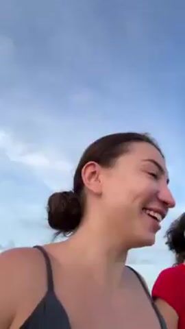 A woman with dark hair pulled back in a bun is smiling and taking a selfie video with another woman. The woman taking the video is wearing a black tank top. The other woman is wearing a red t-shirt and white shorts, with a small purse slung over her shoulder. They are standing on a grassy hill overlooking a body of water and more hills in the distance. The sky is cloudy and the lighting suggests it's dusk. At one point the woman taking the video makes a peace sign.