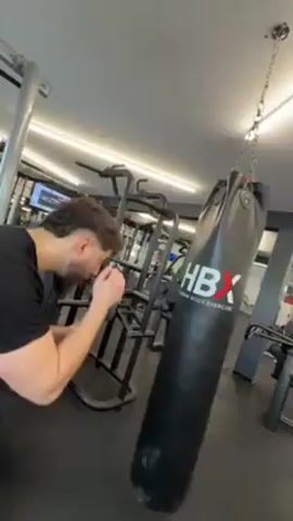 A man with a mullet wearing a black t-shirt and black pants is punching a black punching bag that has the letters "HBX" in white on it. The man is in a gym with gym equipment visible in the background.