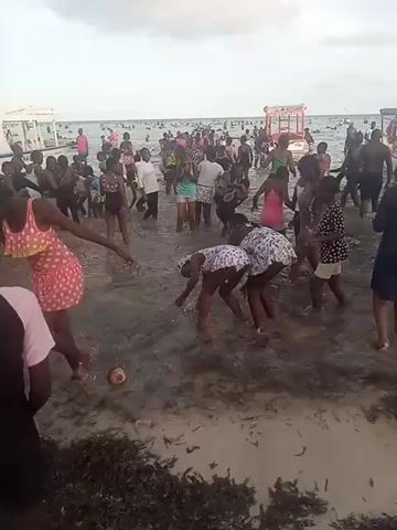 The video shows a crowded beach on a cloudy day. Many people are visible in the water, and a pink, two-story structure can be seen floating in the distance. A group of approximately ten young girls are standing in the shallow water near the shore. They are mostly dressed in swimsuits, with some wearing light-colored dresses or shorts. The girls are laughing and appear to be playfully pushing and splashing each other. The water is murky, and there is a coconut shell on the sand in the foreground.