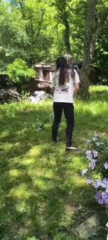 A person with long dark hair, wearing a white t-shirt with a graphic print and black pants, is using a string trimmer to cut grass in a sunny outdoor area with trees and some overgrown vegetation. The person is facing away from the camera and moving from left to right across the frame. A small, weathered building with a red roof is visible in the background, surrounded by trees and foliage. In the foreground, there are some purple flowers. The lighting suggests it is daytime.