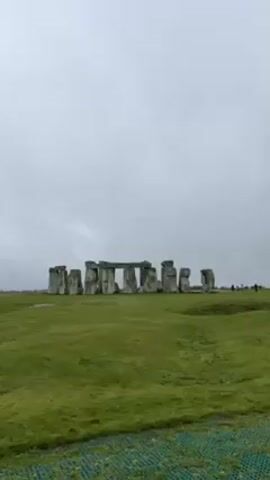The video shows Stonehenge in England on a cloudy day. The monument consists of large upright stones with horizontal stones placed on top. It is surrounded by a green grassy field. There are a few people in the distance. The camera remains still for the duration of the video.