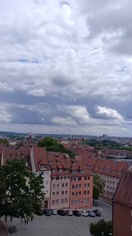 A panning shot reveals a cityscape with terracotta-tiled roofs under a dramatic, cloudy sky. The camera moves across the urban landscape, showcasing rows of buildings with cream-colored facades and dark-tiled roofs. In the distance, a church with twin green spires is visible, along with a sprawling metropolis under a partly cloudy sky. The lower portion of the frame shows a cobblestone courtyard with parked cars.