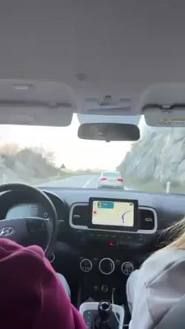 The video shows a view from inside a car driving on a road. The driver's hands are visible on the steering wheel, and a passenger sits in the front seat. The car is following another vehicle, an Audi, and the navigation system on the car's display screen shows a route. In the rearview mirror, the face of the driver wearing a hat can be seen. The surrounding environment includes mountains and roadside vegetation. The weather appears to be clear.