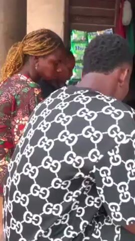 The video shows three people standing outside a small shop. Two of the people are women; one is wearing a colorful patterned top and has blonde braided hair pulled back in a ponytail and the other is wearing a black top. The third person is a man wearing a black and white shirt with Gucci logos. The people are all looking down, possibly at something on the counter of the shop. The shop has some packets of Viva brand detergent visible on the shelves.