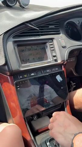 This video shows the interior of a car, focusing on the dashboard and center console. A person's hands are visible, one resting on the gear shift and the other on the touchscreen infotainment system. The infotainment screen displays a reflection of the person in the passenger seat, wearing a white t-shirt with graphics. Another smaller screen, a navigation system, is positioned above the touchscreen. The car's interior features wood-grain trim and some additional gauges above the dashboard. The background seen through the windshield suggests the car is moving, with a blurry landscape of fields and hills. The lighting indicates it is daytime, possibly sunny.