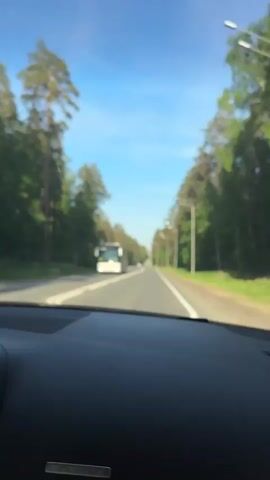 The video shows a view from inside a car driving down a road. The road is straight and has a double yellow line in the center. There are tall trees on both sides of the road. The sky is blue and clear. In the first shot, a bus is passing. Streetlights are visible alongside the road. The car appears to be moving at a moderate speed.