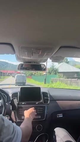 The video shows the interior of a car, looking forward through the windshield. A person's hands are visible on the center console, interacting with the car's media system. The navigation screen is displayed, showing a map with a route. Outside, the car is on a paved road in a rural area with houses and trees. A white van is in front of the car, and other vehicles are further down the road. The sky is blue and sunny. The time stamp progresses from 00:00 to 00:05.