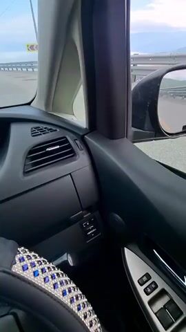 The video begins inside a car, showing the interior of the driver's side, including the steering wheel covered in blue and silver rhinestones, a car door with window controls, and an air vent. The view then shifts to outside the car, looking out the passenger window and through the side mirror. The car is on a road with a yellow line marking the lane. A guardrail runs alongside the road, and beyond it, there are mountains and a hazy sky with clouds. A traffic sign indicating a sharp left turn is visible. The video continues to show the road and the surrounding landscape from the perspective of the moving car.