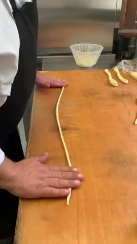 This media file captures a close-up view of a person's hands working on a wooden surface, likely a kitchen counter or workbench. The person, wearing a white shirt and a dark apron, is in the process of shaping a long, thin strand of dough. Their hands are skillfully manipulating the dough, rolling and stretching it to create a consistent thickness.

In the background, several objects are visible, suggesting a food preparation setting. A white bowl, possibly containing cheese or another ingredient, sits on the counter to the right. To the left of the bowl, there are a few other pieces of dough that have already been shaped into similar strands. The overall setting appears to be indoors, with a stainless steel surface visible in the far background, which could be part of a kitchen appliance or workspace. The lighting suggests it is daytime.

The activity being depicted is the creation of handmade pasta or a similar dough-based food item. The focused attention of the hands on the dough implies a careful and practiced process. The context provided indicates this is happening in Castiglione d'Orcia, Italy, which could suggest the dough is for a traditional Italian dish. There is no visible text in the media file.