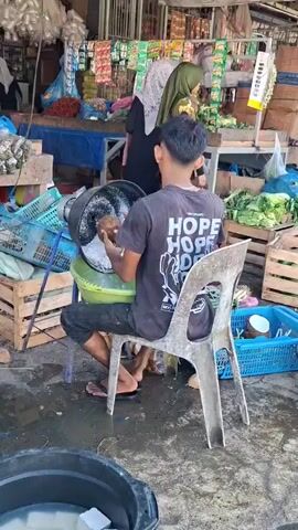 A young man is grating coconut in a busy outdoor market. He sits on a plastic chair, facing away from the camera, and uses a metal grater attached to a black bucket. The grater is being turned by hand, and the grated coconut falls into a yellow basin below. The man is wearing a black t-shirt with the word "HOPE" printed in white on the back, along with a graphic of hands. He is also wearing black pants and flip-flops. In the background, two women wearing hijabs are visible, along with various market stalls selling goods like packaged food items, vegetables, and other produce. There are baskets and crates scattered around the scene, indicating a typical market environment. The ground is wet and appears to be muddy or dirty.