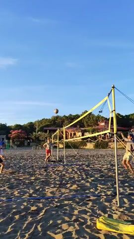 Three people are playing beach volleyball on a sandy beach under a clear blue sky. The person on the left is a female wearing a bikini, positioned low to the ground as if to bump the ball. In the center, a male wearing shorts is jumping up with his arms extended, attempting to hit the ball. To the right, another person, possibly male, is further back and to the side, watching the play. A volleyball net is set up across the sand, and a volleyball is visible in the air. In the background, there are trees, some structures, and playground equipment. The sand is textured with shadows from the lighting, suggesting it is either morning or late afternoon.