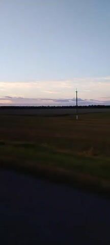 The video shows a view from inside a vehicle, looking out the passenger side window. The vehicle appears to be moving along a paved road. To the right of the road is a grassy area, followed by fields of dry grass and crops. In the distance, there is a treeline and a clear sky with some scattered clouds, indicating either sunrise or sunset. A tall, thin pole, possibly a utility pole or antenna, is visible in the middle ground of the scene. A side mirror of the vehicle is partially visible in the lower left corner of the frame. The overall scene depicts a rural landscape during twilight.