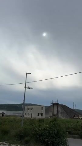 The video shows an airplane approaching for landing under a cloudy sky. The sun is visible, but obscured by the clouds. In the foreground, there's a street lamp, a small building with an air conditioning unit, a weathered concrete structure, and overgrown vegetation. Behind these objects, a mountain range can be seen in the distance, and to the right, a larger industrial building is partially visible. A telephone wire stretches across the top center of the frame. The airplane is green and white, flying relatively low and getting closer.