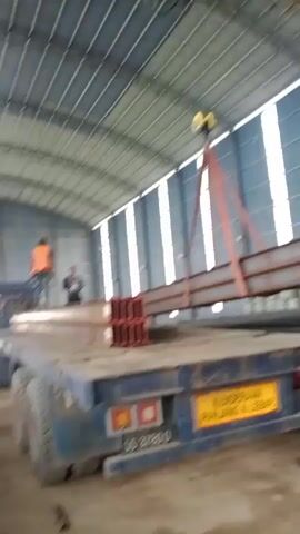 A yellow overhead crane is lifting a long, rusty steel beam with red straps. The beam is being moved from a flatbed truck. Two men are visible on the truck, one wearing an orange vest. The truck has a yellow license plate with black lettering. The scene takes place inside a large, open building with a metal roof and striped walls.
