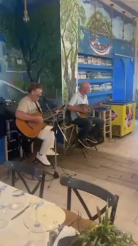Two men are playing music in what appears to be a Greek restaurant. The man on the left is playing a guitar and singing into a microphone. He is wearing a white shirt and white pants. The man on the right is playing a bouzouki. He is wearing a light-colored short-sleeved shirt and dark pants. They are seated on stools in front of a mural that depicts a Greek landscape. Tables set for dining are visible in the foreground.