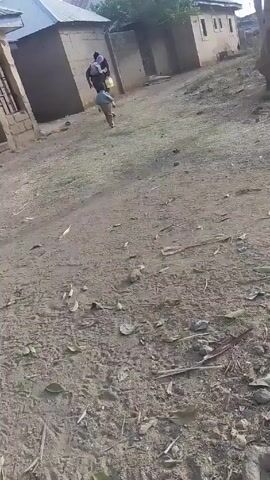 The video shows a woman and a child walking on a dirt path between buildings. The woman is carrying something on her head and the child is walking beside her. The ground is covered in dirt, leaves, and small twigs.  The buildings in the background appear to be simple, one-story structures.