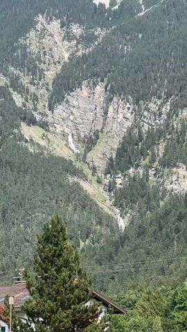 A vertical, zoomed-in video shows a steep, rocky mountain slope covered with dense evergreen trees. Two narrow waterfalls cascade down the gray, layered rock face. The top waterfall is larger and more visible, appearing as a white ribbon against the rock. The lower waterfall is smaller and less distinct. The surrounding mountains are also covered in trees, and a few bare patches of rock are visible. The sky is not visible, but the scene appears to be in daylight. The bottom of the frame shows the tops of trees and a small section of a building with a brown roof.