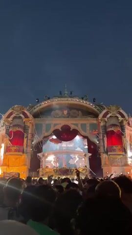 A low-angle, vertical video shows a crowded outdoor music festival stage at dusk. The stage is elaborately decorated with gold and red accents, resembling a grand theater or ballroom. It is illuminated by numerous lights and special effects, including what appears to be pyrotechnics and lasers. A DJ booth is visible on the stage, with a person presumably operating it. The crowd in the foreground is densely packed, with many people looking towards the stage. The sky above is a deep blue, indicating twilight.