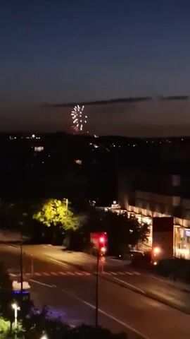 The video shows a nighttime cityscape with fireworks being set off in the distance. In the foreground, a street with a traffic light is visible. The traffic light is red, indicating that traffic should stop. Trees and buildings are also present in the scene. The sky is dark, with a few clouds visible. The fireworks are bright and colorful, and they are creating a spectacular display against the night sky.