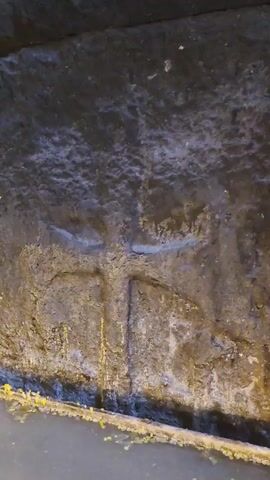 The video shows a close-up of a rough stone wall with a carved cross. Water with melted wax drips from above and collects at the bottom of the frame, where lit candles are floating. The scene appears to be within a dimly lit, possibly ancient structure, with additional carved stone elements and reflections visible in the water.