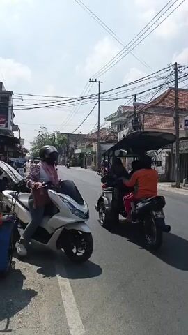 A person wearing a helmet and a pink and white patterned shirt rides a white scooter on the right side of a paved road. The scooter has a black windshield and black accents. The person is also wearing blue jeans and white clogs. The road is lined with buildings on both sides and overhead power lines. There are other vehicles, including motorcycles and a three-wheeled vehicle, parked on the left side of the road. The sky is clear and blue with some clouds. The scene appears to be in a town or city, possibly in Southeast Asia, given the style of the buildings and vehicles.