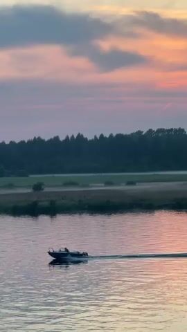 A small motorboat is moving across a calm body of water from left to right. The boat has a dark hull and a white cabin. There are at least two people visible in the boat. The water reflects the pink and orange colors of the sky, which is filled with clouds. In the background, there is a dark treeline on the horizon, with a strip of lighter green fields in front of it. The sky is illuminated by a setting sun, which is partially visible behind the trees, casting a reddish glow. The overall scene depicts a tranquil sunset over a body of water with a boat passing by.