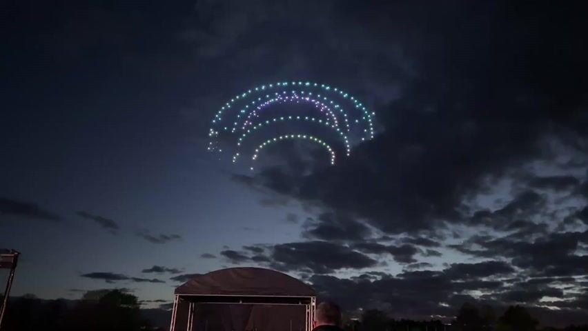 At dusk, a coordinated display of drones, arranged in a three-tiered arc shape, lights up the sky with green, blue, and purple hues. The backdrop is a dark sky filled with scattered clouds. Below, a black canopy tent and silhouette of a person watching the display can be seen. The atmosphere suggests an outdoor event or performance.