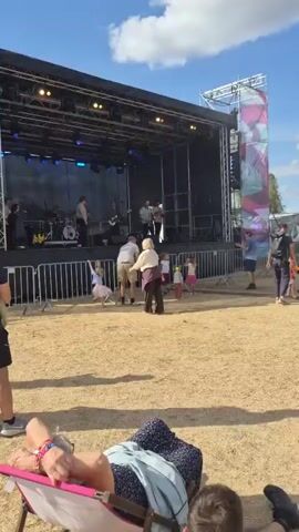 The video shows an outdoor music festival. A band is performing on a stage. The band consists of a guitarist, a bassist, a saxophonist, and a drummer (partially visible). In the foreground, there are several audience members, including children and adults. Some children are dancing, and some adults are standing and watching the performance. The stage is set up outdoors, with a large black backdrop and lighting equipment. There are metal barriers separating the stage from the audience. The ground appears to be covered in sand or dirt. The weather seems to be sunny and warm. The overall atmosphere is festive and relaxed.