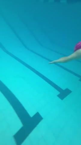 An underwater shot shows a swimmer in a maroon swimsuit with white stripes and a pink swim cap. The swimmer is performing a glide, with arms extended forward and legs together. The swimmer is moving through a bright blue swimming pool with black lane lines visible on the bottom. The shot is slightly tilted, giving a sense of motion and depth.