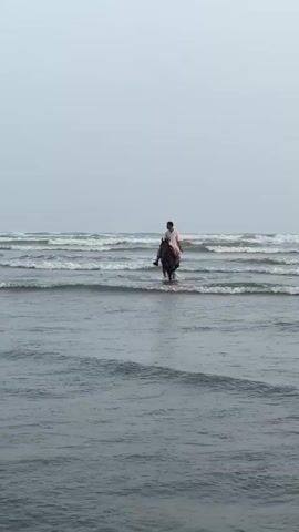 A person rides a horse into the ocean. The rider is wearing a white outfit. The horse is dark brown. The water is choppy, with waves breaking around the horse and rider. The sky is overcast and a light grey. The scene appears to be a beach with a vast expanse of water. The overall mood is calm and peaceful. It is daytime, but the cloudy weather creates a subdued lighting. The location is likely a beach in Karachi Division, Pakistan, given the context. There is no visible text in the media file.