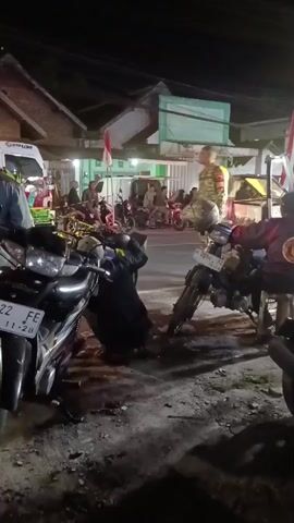 The video shows a nighttime scene on a street, possibly in a rural or suburban area. Several motorcycles are parked or leaning on the ground. In the foreground, a black motorcycle with license plate number 5522 FE is visible. To its right, another motorcycle is also present. A person wearing a dark uniform is crouched down near the motorcycles, appearing to be working on something. A man in a camouflage uniform stands to the right of the crouched person, holding a helmet. Further in the background, there are buildings, some with lights on. A white truck with blue lights on its roof is parked in the middle ground. Several other people are gathered in the background, some seated and others standing. One person in the background is holding a small Indonesian flag. The ground appears to be unpaved and dusty. The lighting suggests it is dark, with artificial light sources illuminating the scene.