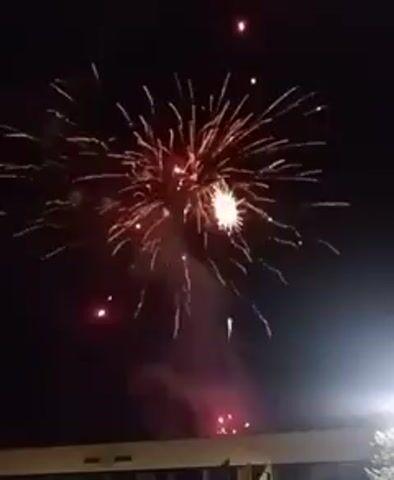 The video captures a nighttime fireworks display. Bright golden-yellow fireworks explode against a dark sky, creating a starry effect. This is followed by a vibrant red explosion, and then another golden-yellow burst. The final explosion is a large, brilliant white one, generating significant smoke and light. Throughout the video, bright lights are visible in the lower right corner, possibly from streetlights or another light source. A dark, elongated structure, possibly a building, is visible at the very bottom of the frame. The event takes place at night, and there are no people or text visible in the video. The user-provided context indicates the location is Yambol, Bulgaria.