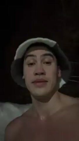 A shirtless young man wearing a hat stands in front of a snowbank in darkness. He rubs handfuls of snow on his face.