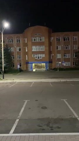 The video begins with a stationary shot of a brick building at night, lit by streetlights. A parking lot with white markings is in the foreground. The camera then pans to the right, showing more of the building, some evergreen trees, and a street with parked cars. The pan continues, revealing a street lamp, a digital billboard, and a street sign. The scene then shifts to a different perspective, showing a sidewalk, a building with glass windows and illuminated signs, a street, and another digital billboard. The video ends with a shot of the street at night, with streetlights and illuminated buildings in the background.