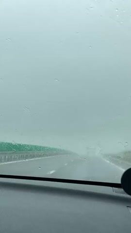 The video shows a first-person view from inside a car driving on a highway during heavy rain or mist, significantly reducing visibility. The car's windshield is covered in water droplets, and the wipers are actively moving to clear the view. The dashboard is visible at the bottom of the frame. In the distance, a large truck can be vaguely seen through the thick fog or rain on the highway. A green barrier or fence runs along the left side of the road. The road surface appears wet. The overall scene suggests challenging driving conditions.