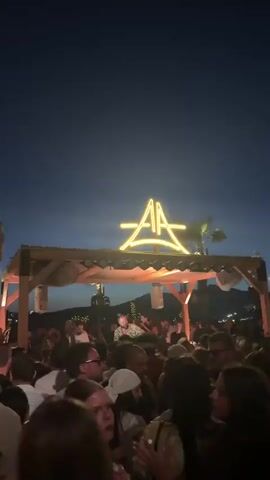 This is a vertical video of an outdoor nighttime party scene. In the foreground, a crowd of people are visible, many with their arms raised. In the background, a DJ is performing on a stage under a wooden canopy. A large neon sign with the letters "AA" is illuminated above the stage. The environment appears to be a beach or a resort area, with palm trees and distant hills visible. The lighting is primarily from stage lights and the neon sign, creating a lively and festive atmosphere. The video transitions between different angles and focus points within the crowd and on the DJ.