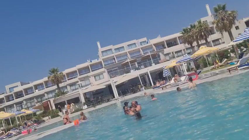 The video opens with a wide-angle shot of a resort swimming pool on a sunny day with a clear blue sky. In the foreground, the bright blue water of the pool occupies most of the frame, with several people partially submerged and interacting. To the left and right of the pool, lounge chairs are visible, some occupied by people, and several umbrellas provide shade. In the background, a large, multi-story resort building dominates the scene, with balconies and windows. Palm trees are scattered around the resort grounds. The camera appears to be positioned low, close to the water's surface, and is tilted at an angle, giving a dynamic perspective. The overall impression is a lively vacation scene.