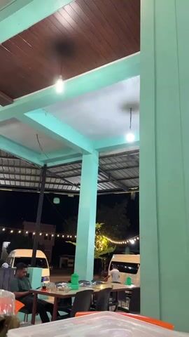 This media file captures an outdoor seating area at night, likely a restaurant or food stall, in Grong-Grong, Indonesia. The setting features a covered structure with teal-painted pillars and beams, supporting a tin roof and a wooden ceiling. Exposed light bulbs hang from the ceiling, illuminating the space.

In the foreground, a white table is visible with various objects, including a clear plastic container, a pink packet of "Jolly" tissues, and a glass containing a dark liquid, possibly tea. Behind this table, another table is set up with chairs. A man is seated at this table, looking down at his phone. He is wearing a dark green t-shirt. A green bottle and some food items are also on this table.

Further into the background, more tables and chairs are arranged. Two white vans are parked, and string lights are strung across the area, creating a festive ambiance. The overall scene suggests a relaxed, casual dining experience in the evening. The time of day is clearly night, indicated by the dark sky and artificial lighting. There are no immediate weather cues, but the covered structure suggests protection from potential rain or sun. The presence of food, drinks, and people suggests a social gathering or a meal. The visible text "Jolly" on the tissue packet is the only discernible text.