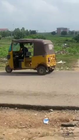 The video shows a yellow tuk-tuk, a three-wheeled vehicle often used as a taxi, driving slowly on a paved road. The tuk-tuk has a brown canvas roof and appears somewhat worn. A man is driving the vehicle, wearing a black shirt and black pants. The background includes green vegetation, scattered debris, and buildings in the distance. The sky is slightly overcast.